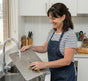 A happy female customer holding a titanium chopping board under running water in a kitchen sink. The board rinses clean instantly without scrubbing, demonstrating the easy-to-clean and non-stick benefits of titanium.