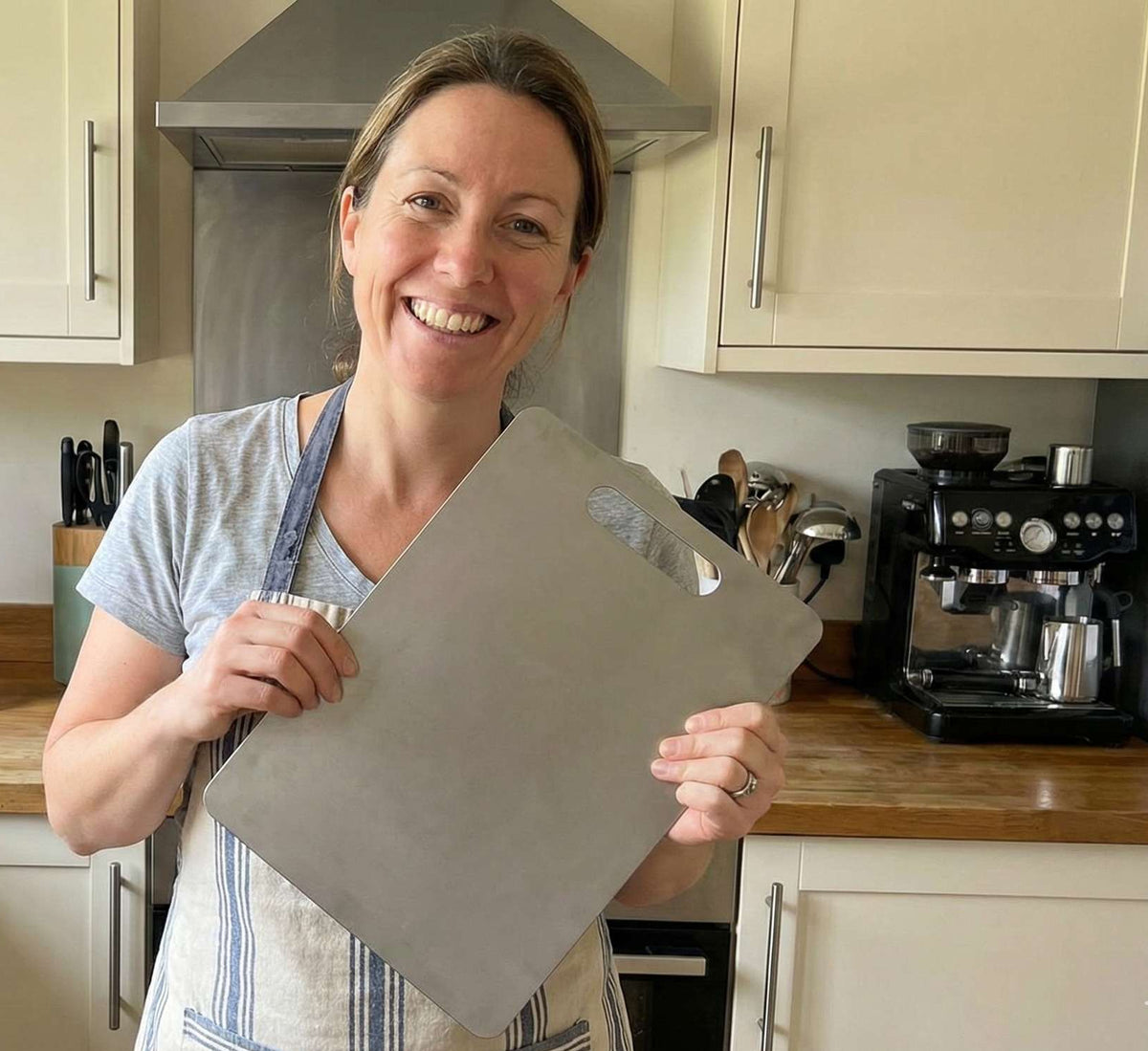 Smiling woman in her home kitchen holding her Life Upgrade Co titanium chopping board, sharing a 5-star product review.